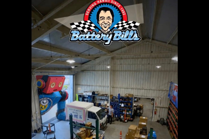A high-angle view of a warehouse interior with the Battery Bills logo prominently displayed at the top. The space contains shelving, boxes, equipment, and a large blue and red cartoon mural on the left wall.